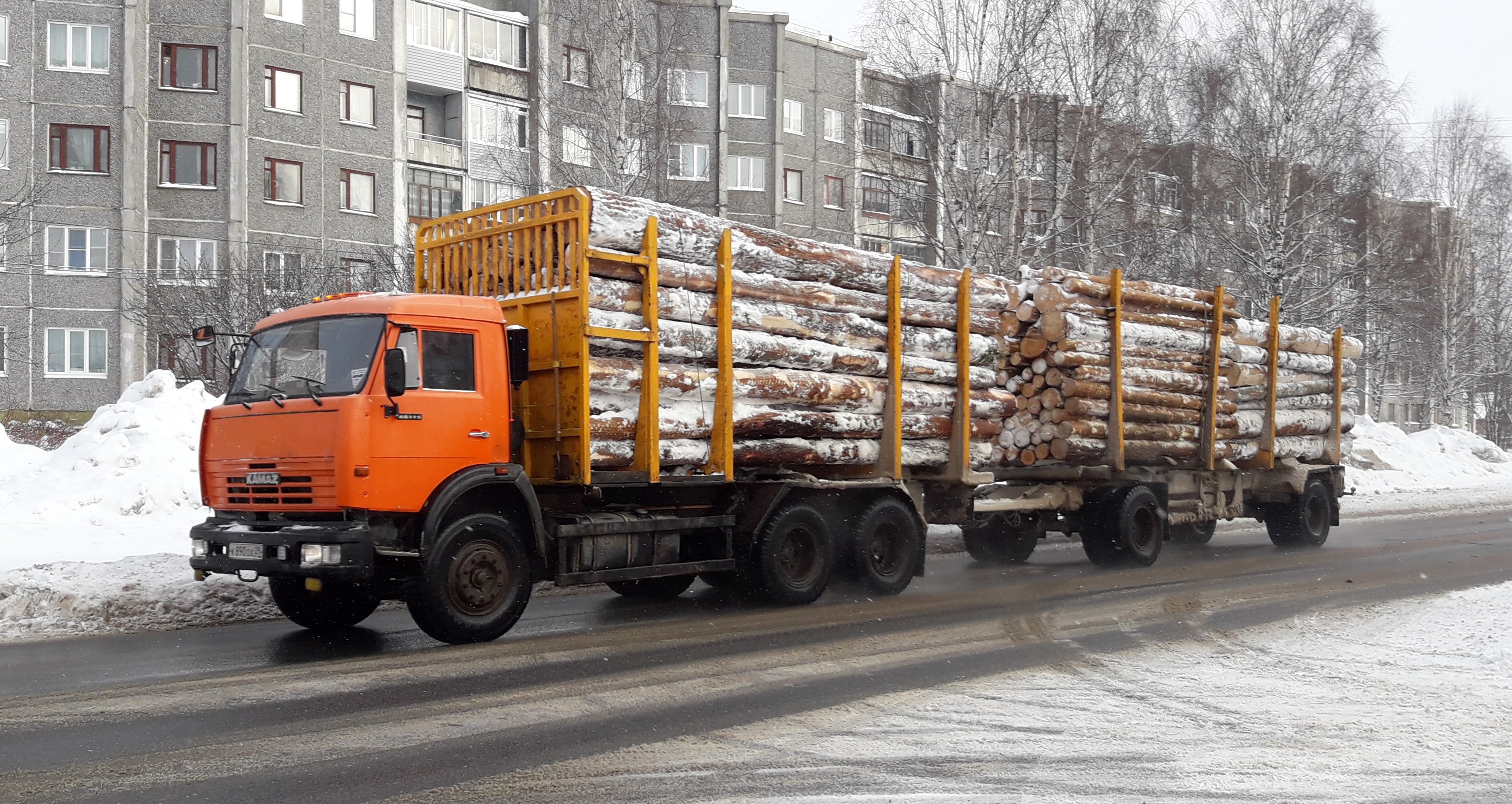 Ein Lastwagen mit Holz fährt auf einer verschneiten Straße mit Bäumen, Gebäuden und einem klaren Himmel im Hintergrund.