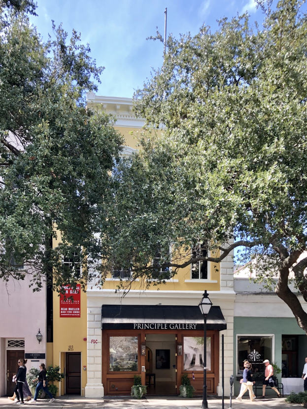 Gruppe von Menschen, die auf einem Gehweg vor dem Principle Gallery Building in Charleston, SC, mit Topfpflanzen, einer Straßenlaterne, einem Schild, einer Fahne, einem Fahnenmast und einem bewölkten Himmel gehen.
