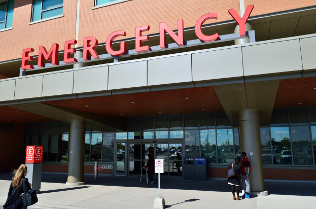 Eine Gruppe von Menschen vor einem Glaswand-Notfallgebäude mit Säulen, einem Namensschild, Hinweistafeln auf Pfählen und einem Mülleimer.