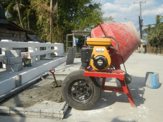 Betonmischer auf einem Zementlaster umgeben von Baustoffen und einer Wohngegend unter einem bewölktem Himmel.