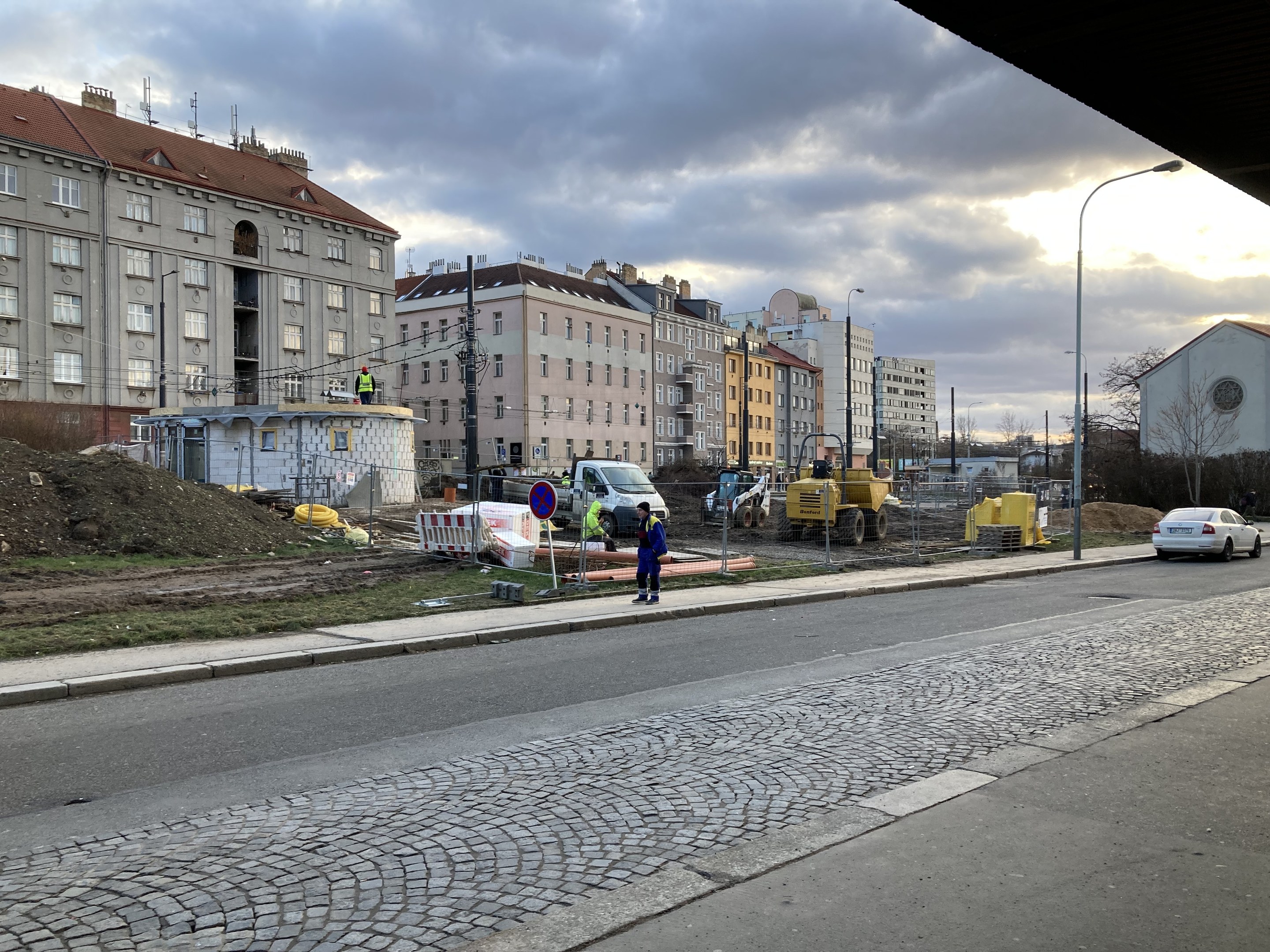 Stadtstraße mit parkenden Autos, Gebäuden, Laternen, Bäumen und einem bewölktem Himmel, mit einer Sperre mit Fahrzeugen und Füßgängern im Vordergrund.