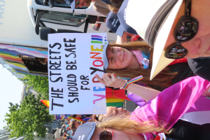 Eine Gruppe von Menschen steht vor einem Lkw und hält ein Schild hoch, auf dem "Die Straßen sollten sicher sein". Einige der Menschen tragen Mützen und Schutzbrillen, andere halten Schilder. Im Hintergrund gibt es Bäume, Gebäude und einen klaren blauen Himmel.