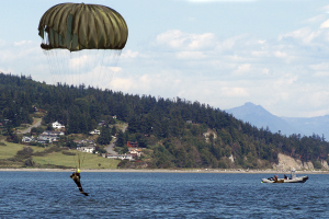 Eine Person gleitschirmfliegt über Wasser mit einem Boot auf der rechten Seite, Bäume, Gebäude, Hügel und einen klaren blauen Himmel im Hintergrund.