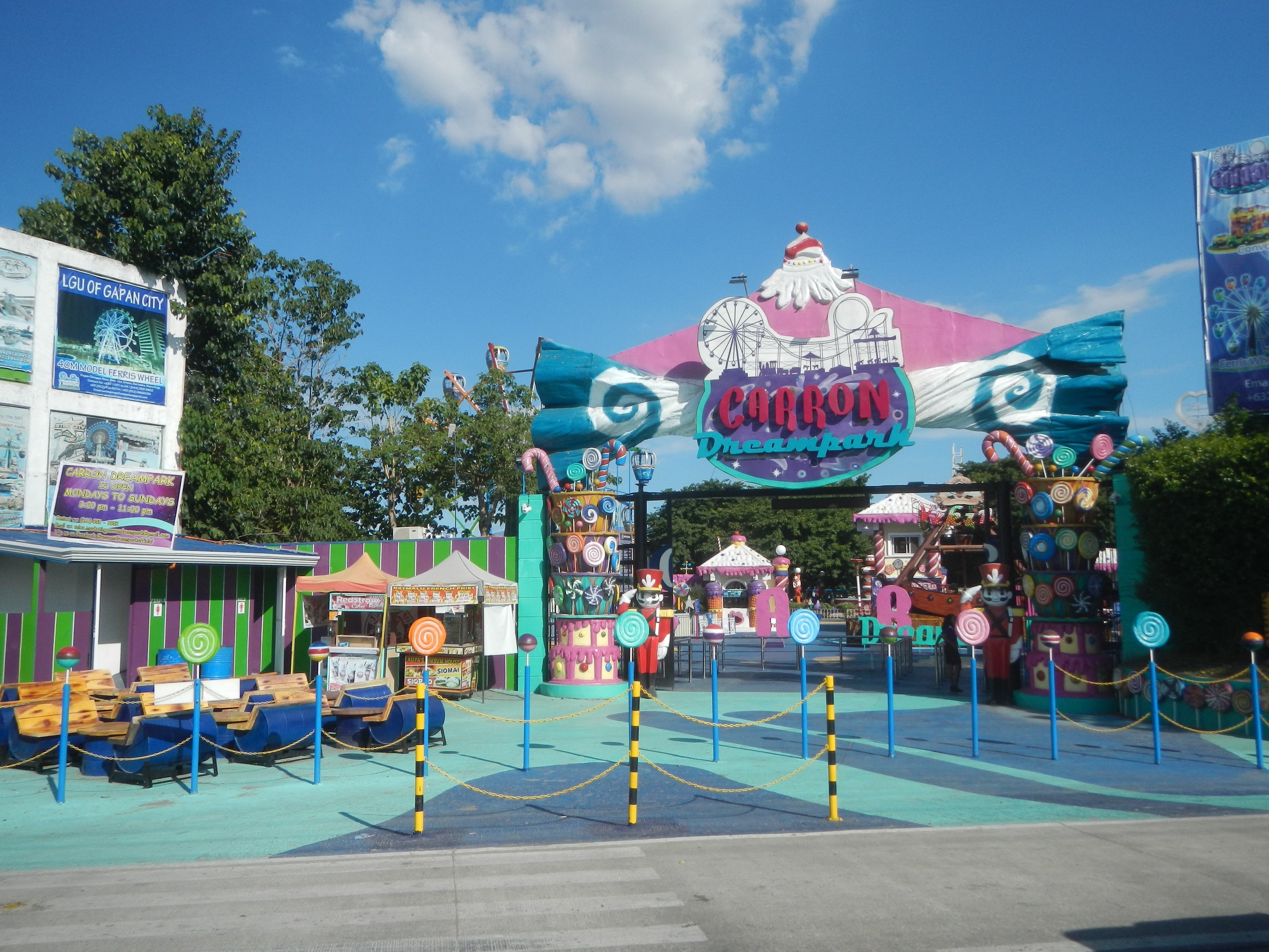 Kirmesfahrgeschäft am Parkeingang mit Straße darunter, eingezäuntem Bereich, Buden, Banner, Bäumen und bewölktem Himmel im Hintergrund.