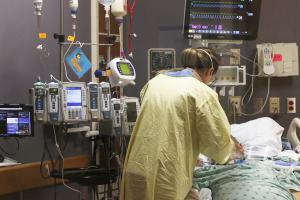 Ein Mann liegt in einem Krankenhausbett neben einem Monitor, mit einer Frau neben ihm; ein Tisch mit einem Monitor ist links, und medizinische Geräte sind im Hintergrund.