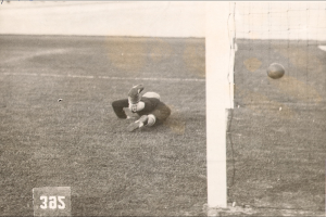 Ein Schwarz-Weiß-Foto eines Fußballspielers, der einen Ball vor einem Tor mit Netz auf der rechten Seite und Text unten fängt.