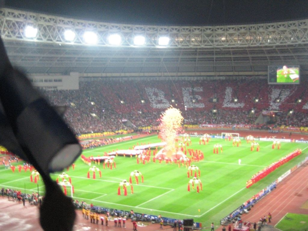 Ein Stadion mit Flutlicht, Eisengittern, Zuschauern, stehenden Personen auf dem Boden, Werbetafeln, Bildschirmen, einer Überwachungskamera, Fahnen und Dekorationen.