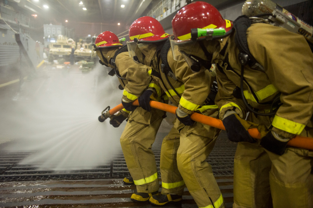 Feuerwehrleute in Helmen und Handschuhen sprühen Wasser aus Rohren auf ein Feuerwehrauto, mit verschiedenen Gegenständen und Texttafeln im Hintergrund und einem Boden unten.