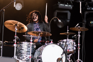 Ein Mann mit Dreadlocks, der auf der Bühne Schlagzeug spielt und Drumsticks hält, mit Mikrofonstativen vor ihm, vor dunklem Hintergrund mit Bühnenbeleuchtung.
