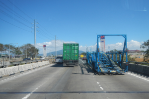 Ein Lastwagen mit einem weiteren großen Lastwagen auf der Autobahn unterwegs, mit Leitplanken, Strommasten, Laternen, Bäumen, Gebäuden und einem bewölkten Himmel im Hintergrund.