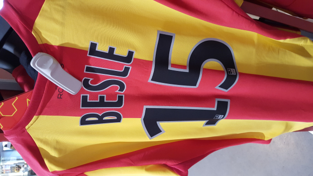 Rotes und gelbes Fußballtrikot mit Text und Zahlen, das auf einem Ständer in einem Geschäft hängt, mit anderen Gegenständen und dem Boden im Hintergrund.