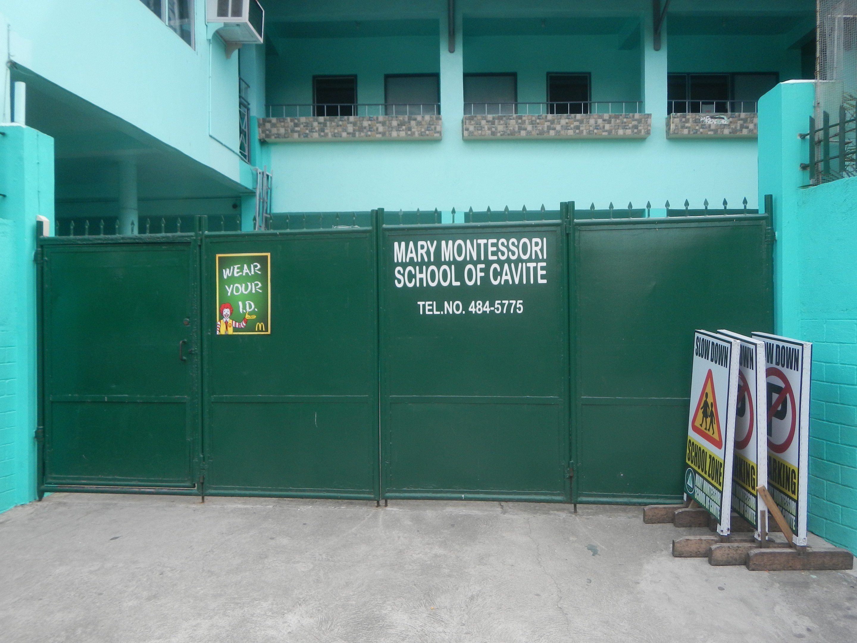 Mary Montessori School of Cavite, a multi-story building with windows, railings, pillars, and air conditioners, featuring a green gate with a sign and boards with text on the ground to the right.