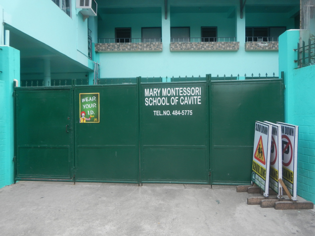 Mary Montessori School of Cavite, a multi-story building with windows, railings, pillars, and air conditioners, featuring a green gate with a sign and boards with text on the ground to the right.