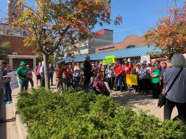 Eine Gruppe von Studenten protestiert vor einem Gebäude der University of Michigan, hält Schilder und einige tragen Kameras und Taschen, mit Bäumen und Himmel im Hintergrund.