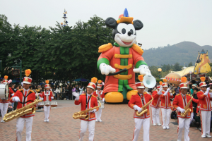 Eine Gruppe von Menschen in Kostümen, die Instrumente spielen, vor einem Mickey-Maus-Floß während eines Umzugs, mit Bäumen, Gebäuden, Hügeln und einem klaren blauen Himmel im Hintergrund und einer Tafel mit Text links.