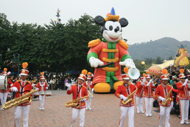 Eine Gruppe von Menschen in Kostümen, die Instrumente spielen, vor einem Mickey-Maus-Floß während eines Umzugs, mit Bäumen, Gebäuden, Hügeln und einem klaren blauen Himmel im Hintergrund und einer Tafel mit Text links.