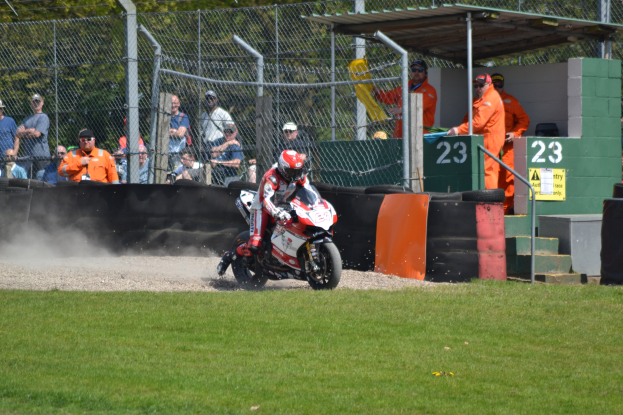 Eine Person in Helm und Handschuhen auf einem Motorrad auf einem grasbewachsenen Feld, mit Zuschauern in Mützen und Schutzbrillen, Maschendrahtzaun, Treppe mit Geländer und einem nummerierten Schuppen im Hintergrund.