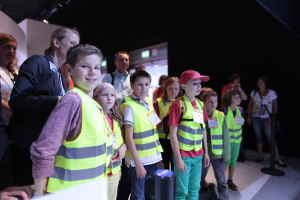 Eine Gruppe von Kindern in Sicherheitwesten und Namensschildern, mit einem Jungen auf der linken Seite, der lächelt, auf einem Boden mit einem Pfahl auf der rechten Seite und ein paar Menschen im Hintergrund.