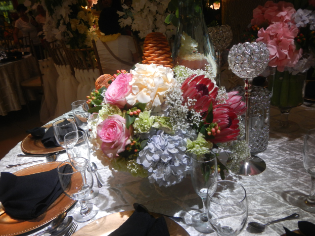Ein formeller Esstisch mit Tellern, Besteck, Gläsern, Blumenvasen und Servietten, mit einem Blumenarrangement in der Mitte, im Hintergrund Stühle und Tische sowie einige sitzende Personen.