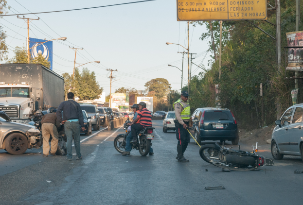 Eine Gruppe von Menschen um ein verunglücktes Motorrad auf der Straße herumsteht, umgeben von mehreren Fahrzeugen, einschließlich eines Lastwagens, und einem Hintergrund aus Bäumen, Pfählen, Lampen und Schildern.