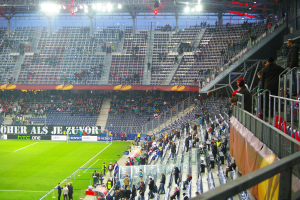 Ein großes Stadion voller Zuschauer, die ein Fußballspiel verfolgen, mit einigen Personen, die auf Stühlen auf dem Feld stehen oder sitzen, einem Geländer und einer Fahne auf der rechten Seite und einem leeren Stadion mit Deckenbeleuchtung im Hintergrund.