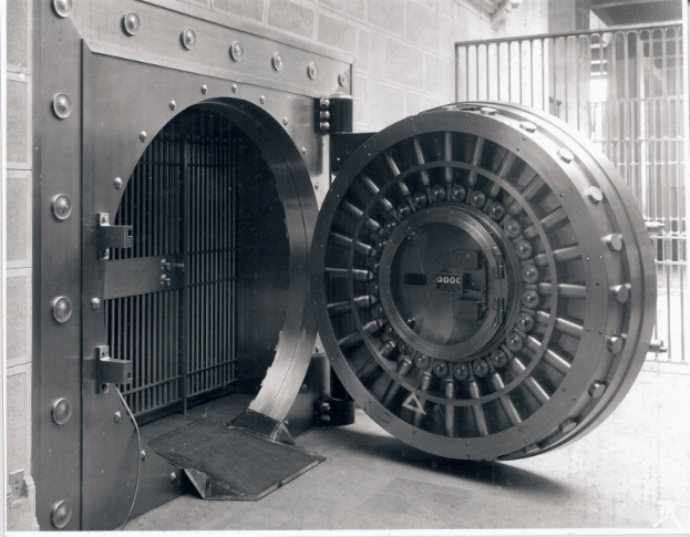 Eine große Metallbanktresortür in einem Gebäude, mit einer Wand und einem Gitter im Hintergrund, in Schwarz-Weiß dargestellt.