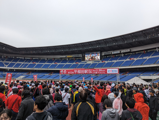 Eine große Menschenmenge steht vor einem Stadion während der Eröffnungsfeier der Olympischen Spiele Tokyo 2020, mit Bannern, Stühlen und einem Bildschirm im Hintergrund bei sichtbarem Himmel.