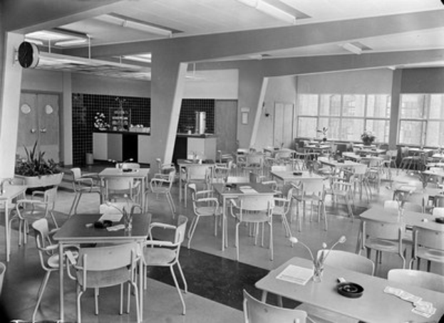 Schwarzes und weißes Foto einer Cafeteria mit Tischen und Stühlen, Tellern, Besteck, Gläsern, Blumenvasen und Deckenleuchten, mit Türen und Fenstern im Hintergrund.