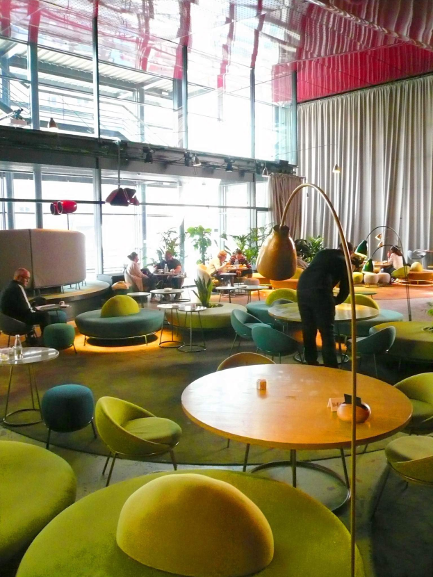 Ein geräumiges Hotel-Foyer mit grünen Stühlen, Tischen, Sofas, stehenden Menschen, Pflanzen, einer Stehlampe, Vorhängen, Deckenleuchten und einer gemütlichen Atmosphäre.
