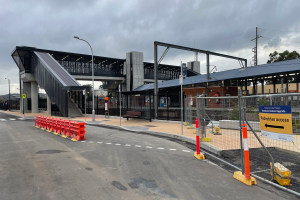 Ein Bahnhof mit einer Brücke im Hintergrund, Straßenlaternen, Strommasten, Stromkabel, Bäume, Gebäude, Fahrzeuge auf der Straße, Absperrpoller, Schilder, Bänke und eine bewölkte Himmel.