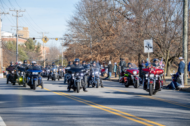 Eine Gruppe von Menschen fährt Motorräder auf einer Straße mit Strommasten, Werbetafeln, Bäumen und Gebäuden unter einem klaren blauen Himmel, wobei einige Fahrer Helme tragen.