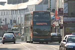 Eine Straße mit Autos und einem Bus vor Gebäuden mit Wänden, Fenstern, Schalen und Döchern, mit Plakaten und Bannern an den Wänden und einem Pfahl mit einer Straßleuchte.