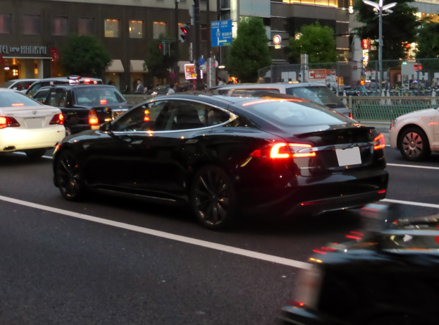 Schwarzer Tesla Model S fährt nachts auf einer Stadtstraße mit anderen Fahrzeugen, Gebäuden, Bäumen, Laternen, Verkehrszeichen, Geländern und Fußgängern im Hintergrund.