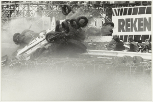 Ein Schwarz-Weiß-Foto eines demolierten Rennwagens, umgeben von Reifen, mit einer Gruppe von Menschen rechts und einer Tafel mit Text im Hintergrund.