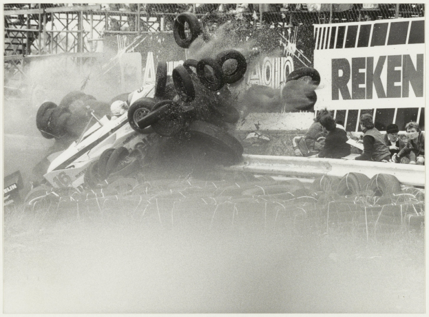 Ein Schwarz-Weiß-Foto eines demolierten Rennwagens, umgeben von Reifen, mit einer Gruppe von Menschen rechts und einer Tafel mit Text im Hintergrund.