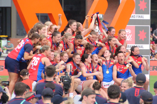 Group of women's AFL players celebrating with a trophy in front of a cheering crowd, some wearing medals and caps, others holding cameras, with boards and a building in the background.