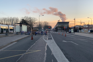 Eine Straßenansicht mit Verkehrskegeln, einem Bus, Laternen, Bäumen, Gebäuden, Kränen und Rauch, der aus einem fernen Gebäude aufklaren Himmel aufsteigt.