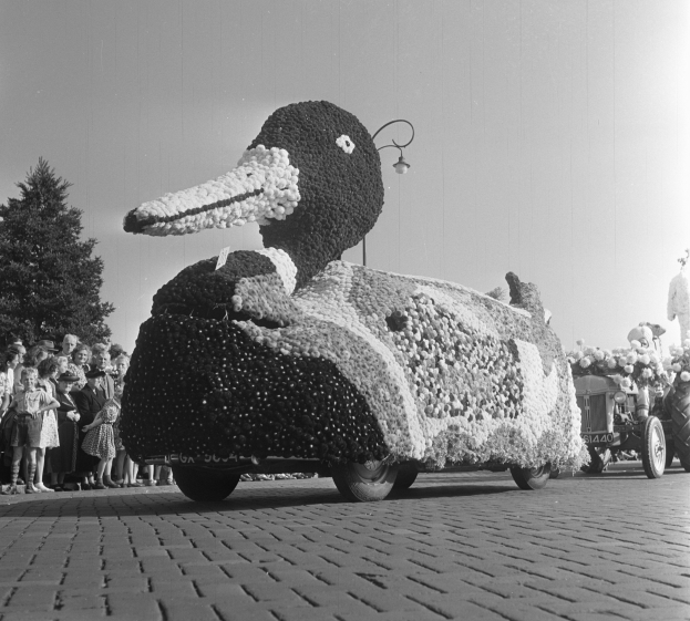 Ein Schwarz-Weiß-Foto eines Entenwagens bei einem Umzug, geschmückt mit Blumen, mit einer Gruppe von Menschen auf der linken Seite, Bäumen und einem klaren Himmel im Hintergrund.