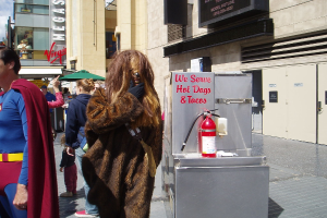 Eine Menge Menschen, einige in Kostümen, versammelt sich vor einem Gebäude mit sichtbarer Beschriftung und Türen, mit einer Kiste und einem Feuerlöscher in der Nähe.