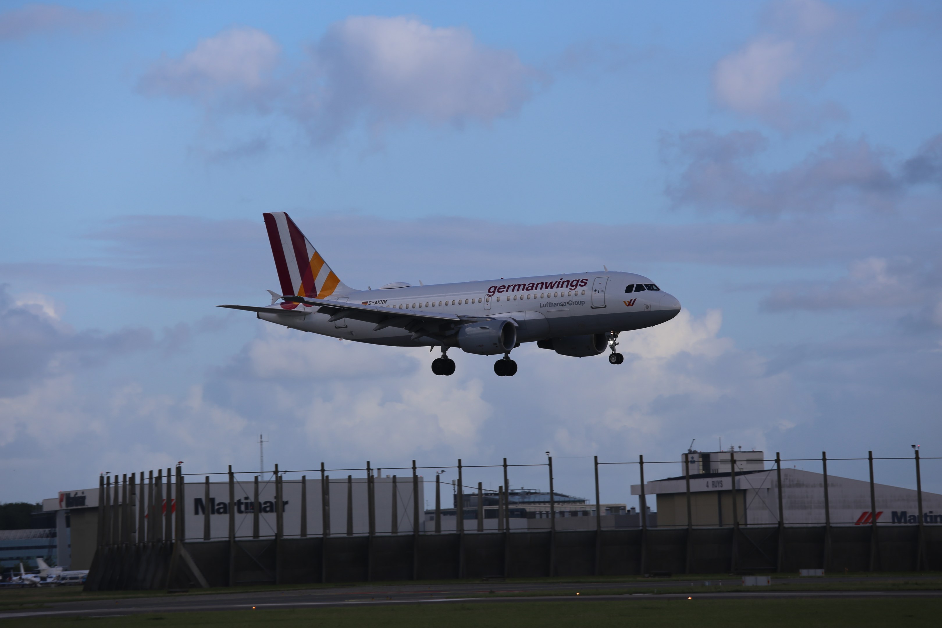 Germanwings Airbus A320-200 beim Start von Frankfurt Airport mit Gras darunter, einem Zaun im Vordergrund, Gebäuden und Bäumen im Hintergrund und einer bewölkten Himmel über