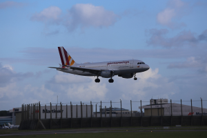 Germanwings Airbus A320-200 beim Start von Frankfurt Airport mit Gras darunter, einem Zaun im Vordergrund, Gebäuden und Bäumen im Hintergrund und einer bewölkten Himmel über