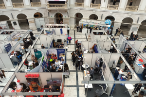 Eine große Gruppe von Menschen auf einer internationalen Fachmesse mit Ständen, Bannern, Tischen, Stühlen, Ballons und einem Gebäude mit Bögen, Säulen, Geländern und einem Bildschirm im Hintergrund.