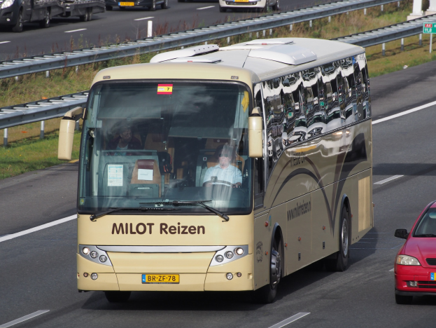 Ein Minibus mit Passagieren auf einer Autobahn neben einem roten Auto fahrend, mit Geländern und Gras an den Seiten und Säulen und Schildern mit Text im Hintergrund.