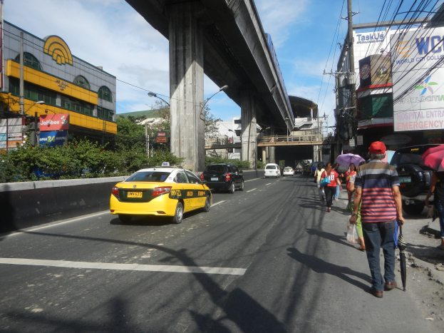Ein gelbes Taxi fährt auf einer Stadtstraße mit hohen Gebäuden, Fußgängern auf dem Gehweg mit Regenschirmen, einer Brücke im Hintergrund, bewölktem Himmel und Oberleitungen.