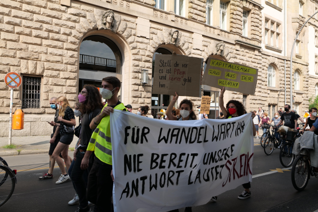 Eine Gruppe von Menschen geht während einer Demonstration die Straße entlang, einige halten Schilder und Banner, andere fahren Fahrräder, im Hintergrund ein Gebäude mit Fenstern, Bögen, Säulen und Skulpturen.