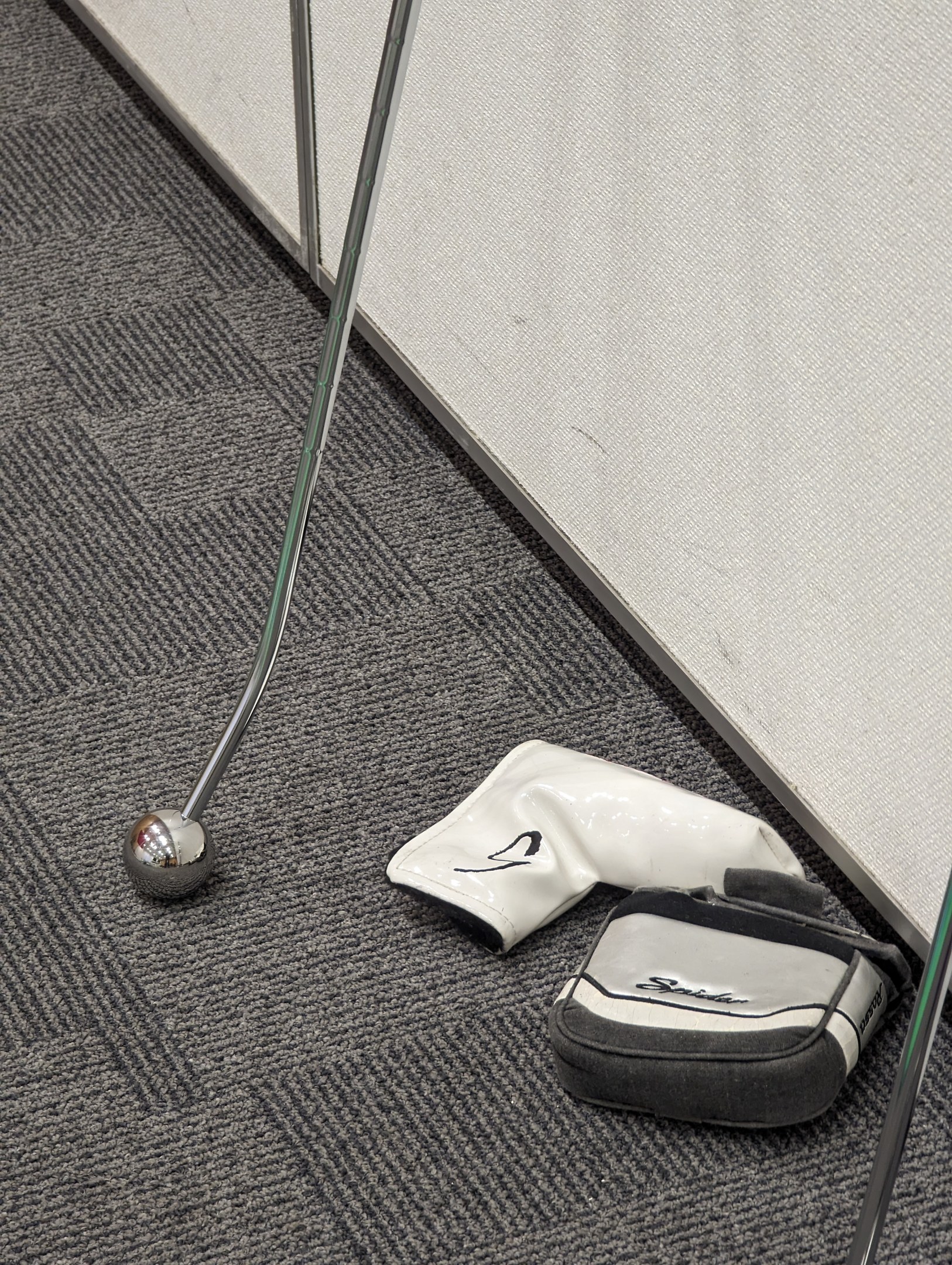 Ein silberner Golfschläger liegt auf einem Teppichboden in der Nähe einer schwarzen Golftasche mit einem weißen Logo, einem Golfschläger und einer hellgrauen Wand mit sichtbaren Kratzern und Dellen.