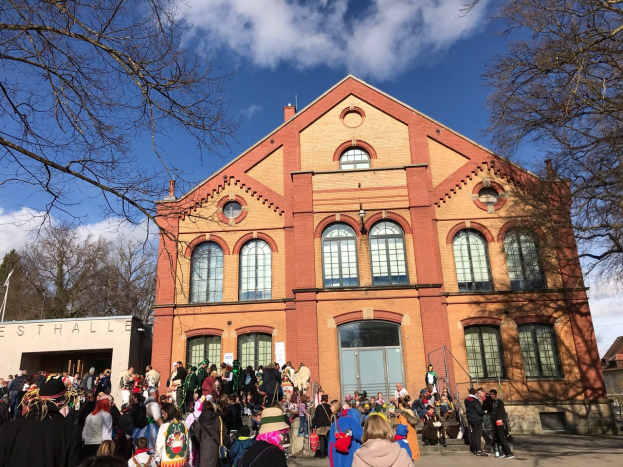 Eine große Gruppe von Menschen steht vor einem mehrstöckigen Gebäude mit Fenstern und Türen, umgeben von Bäumen unter einem bewölkten Himmel, was auf ein Stadtfest hinweist.