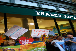 Eine Gruppe von Menschen steht vor einem Geschäft und hält Schilder und Plakate hoch, hinter ihnen ist eine Säule und eine Dachbeleuchtung zu sehen.