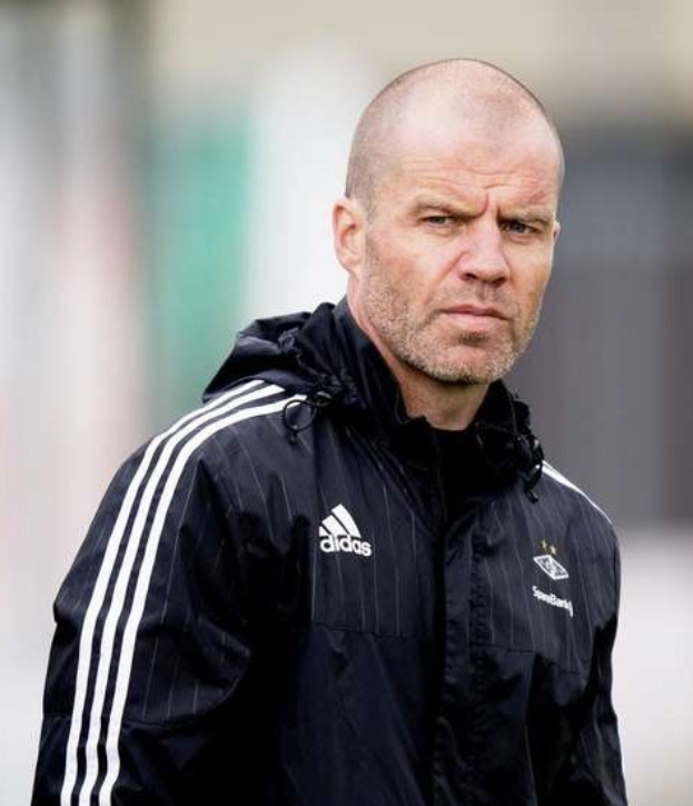 Mann in einem blauen Jackett mit weißen Streifen und weißem Kragen, der auf einem Fußballfeld steht und nachdenklich schaut.