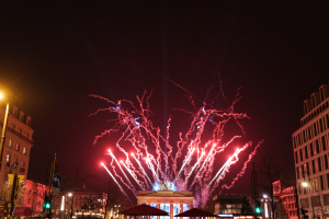 Eine belebte Stadtstraße in Berlin an Silvester, voller Menschen, Fahrzeuge und Gebäude, beleuchtet von Feuerwerk und Lichtern, die eine festliche Atmosphäre schaffen.